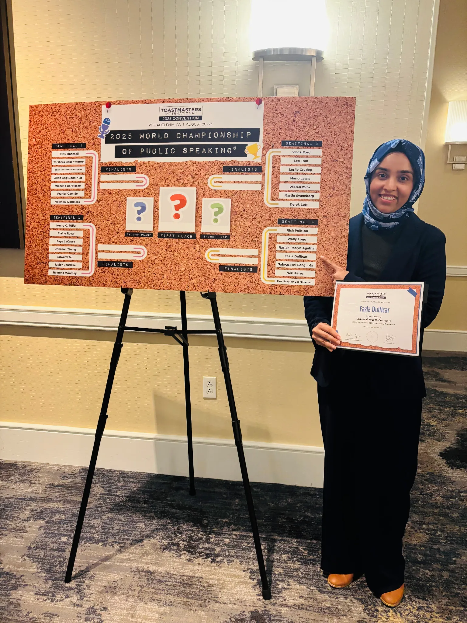 Fazla Dulficar holding her semifinalist certificate beside the 2025 World Championship of Public Speaking board